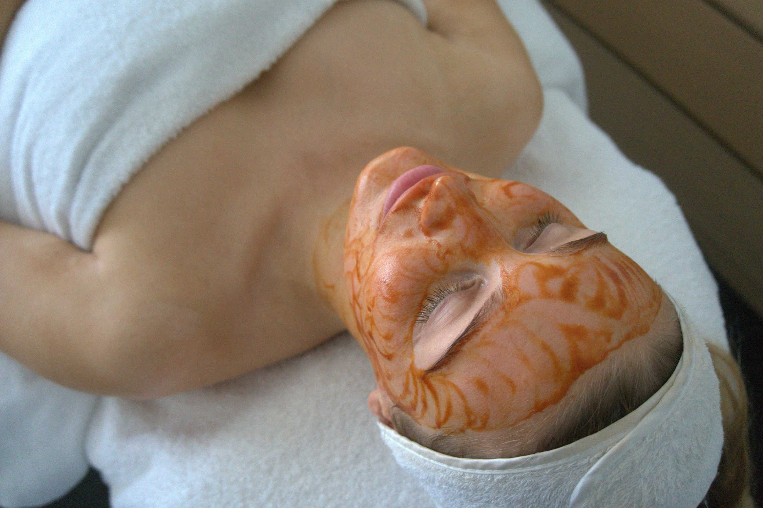 Facial Infusion; A woman lying down with her eyes closed, receiving a facial treatment. Her face has orange and yellow marbled facial mask, and she is covered with a white towel.