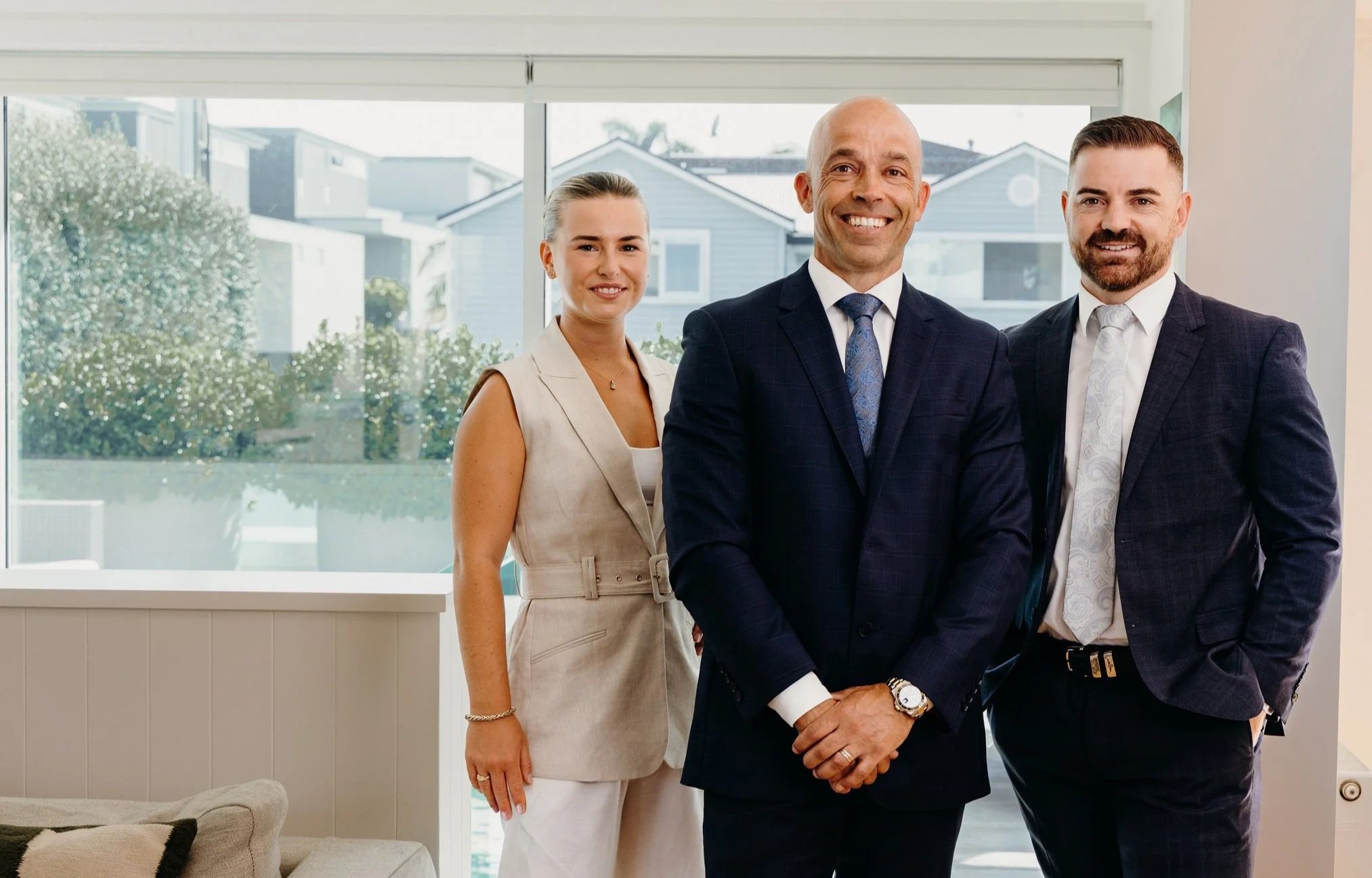 Three professionally dressed people standing indoors in front of large windows, smiling at the camera.