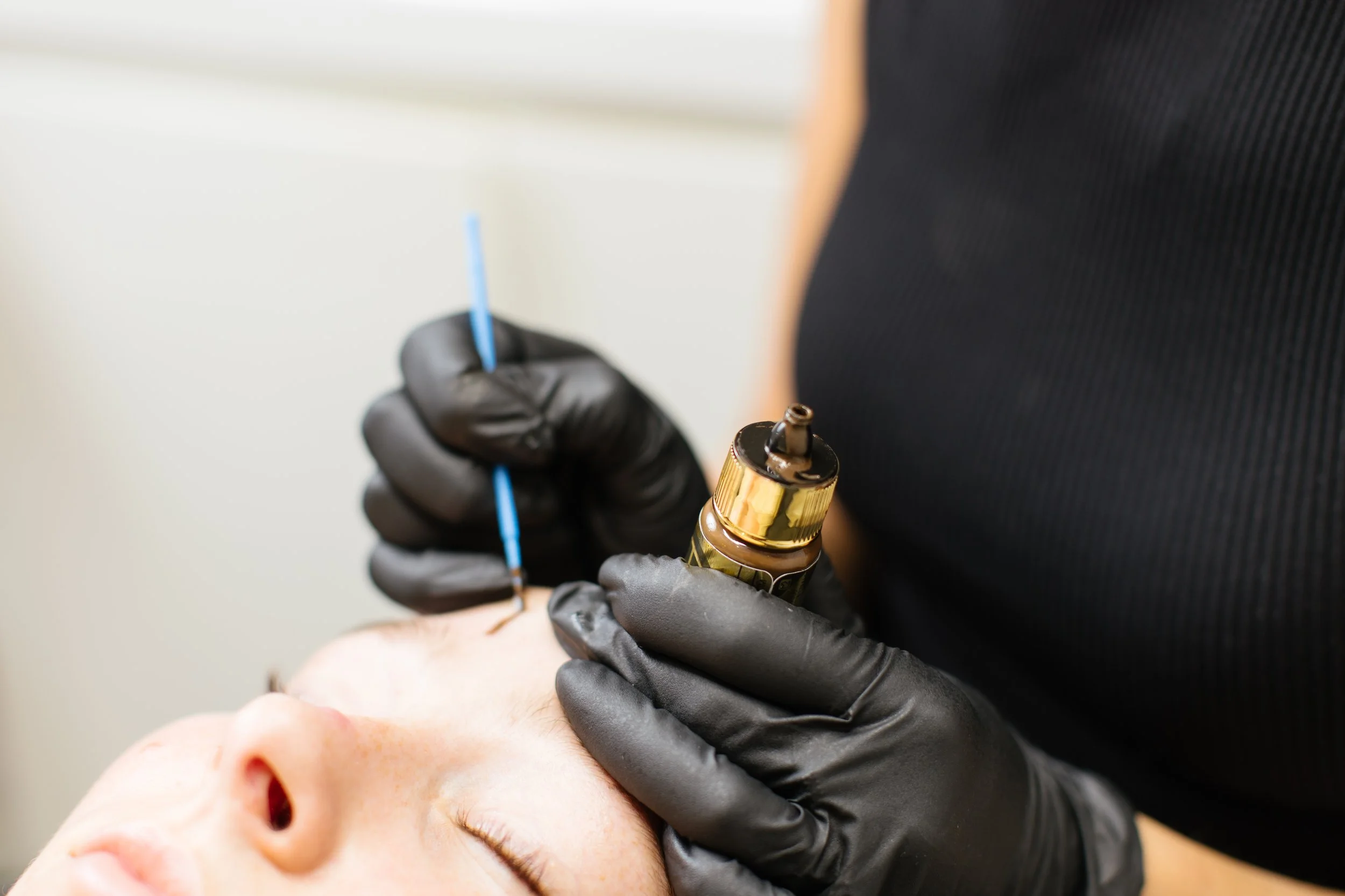 A person lying down receiving a cosmetic microblading procedure. The technician, wearing black gloves, uses a small tool with a blue handle to apply pigment to the client's eyebrows. The client has fair skin and closed eyes.
