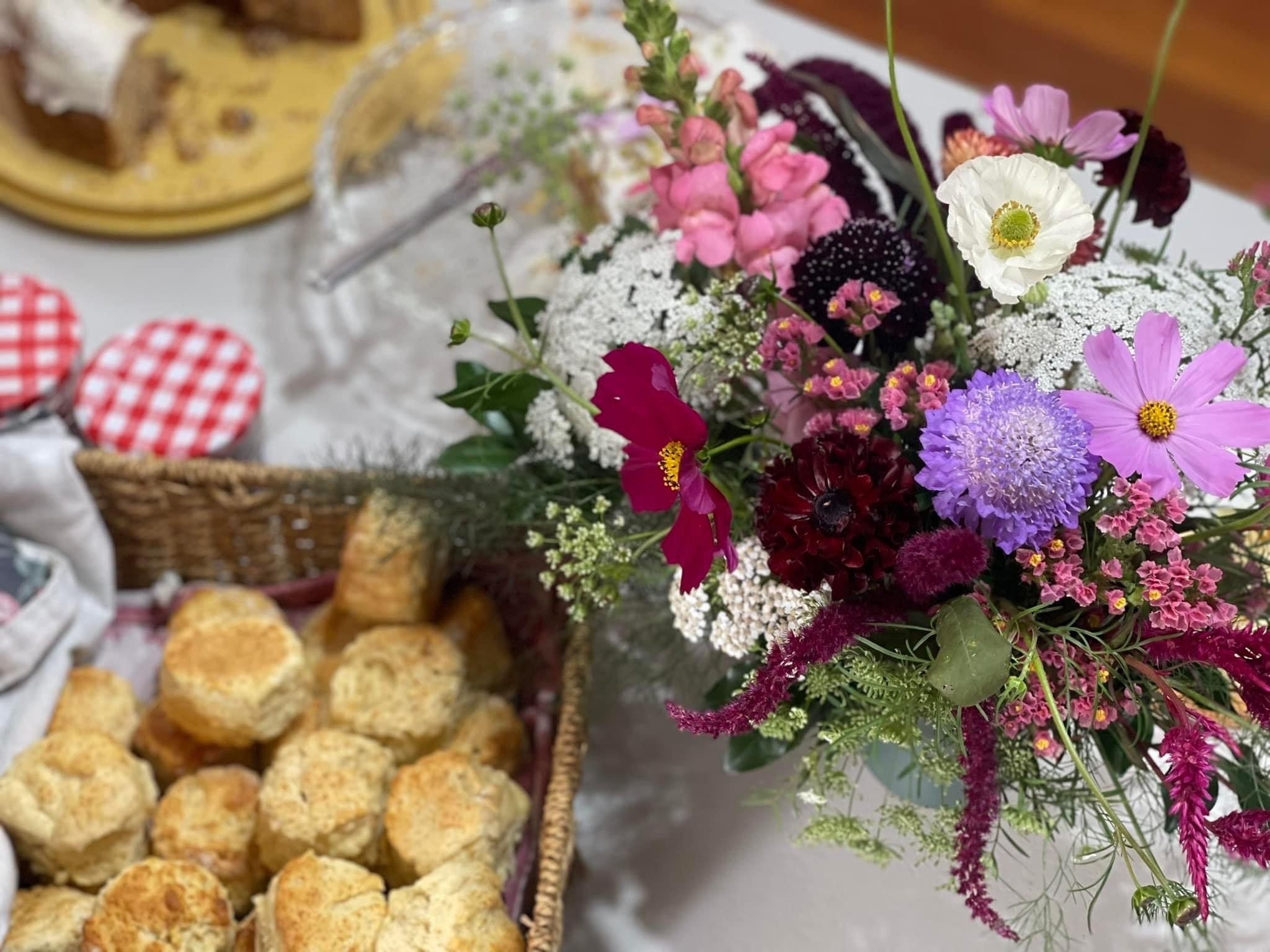 A colorful bouquet of various flowers, including purple, pink, white, and red blooms, next to a basket of scones and a plate of cookies with red and white checkered lids on a table.