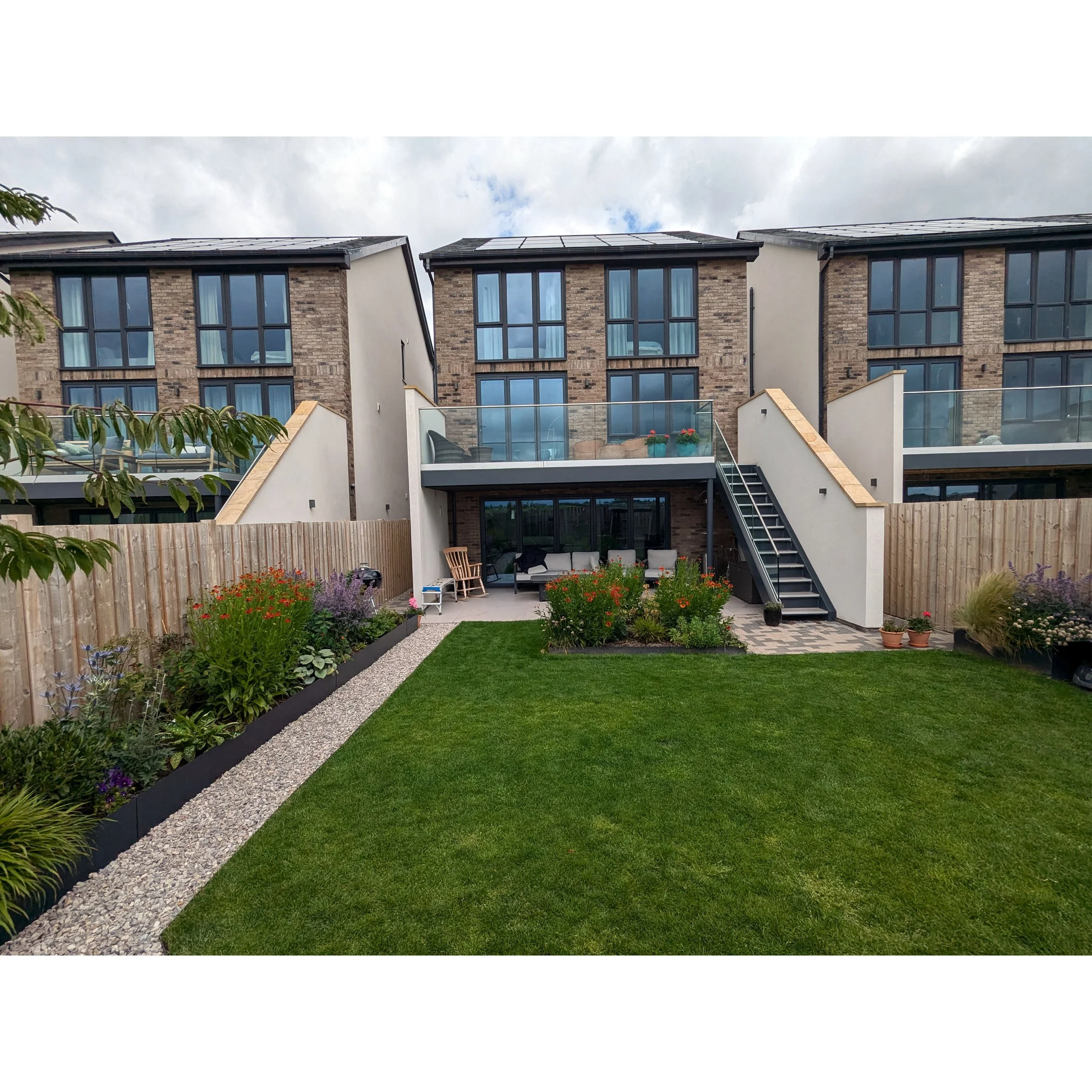 A contemporary townhouse with large windows and a cozy back garden with flowers, grass, and a small paved patio.