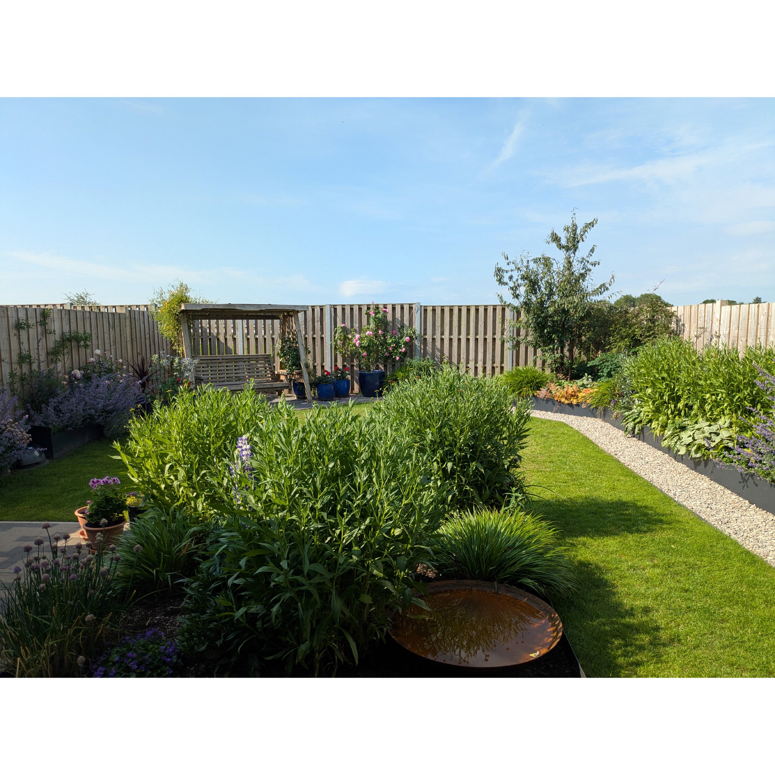 A contemporary back garden with a raised metal planters containing colourful flowers, a garden swing, potted plants, and a gravel pathway under a blue sky.