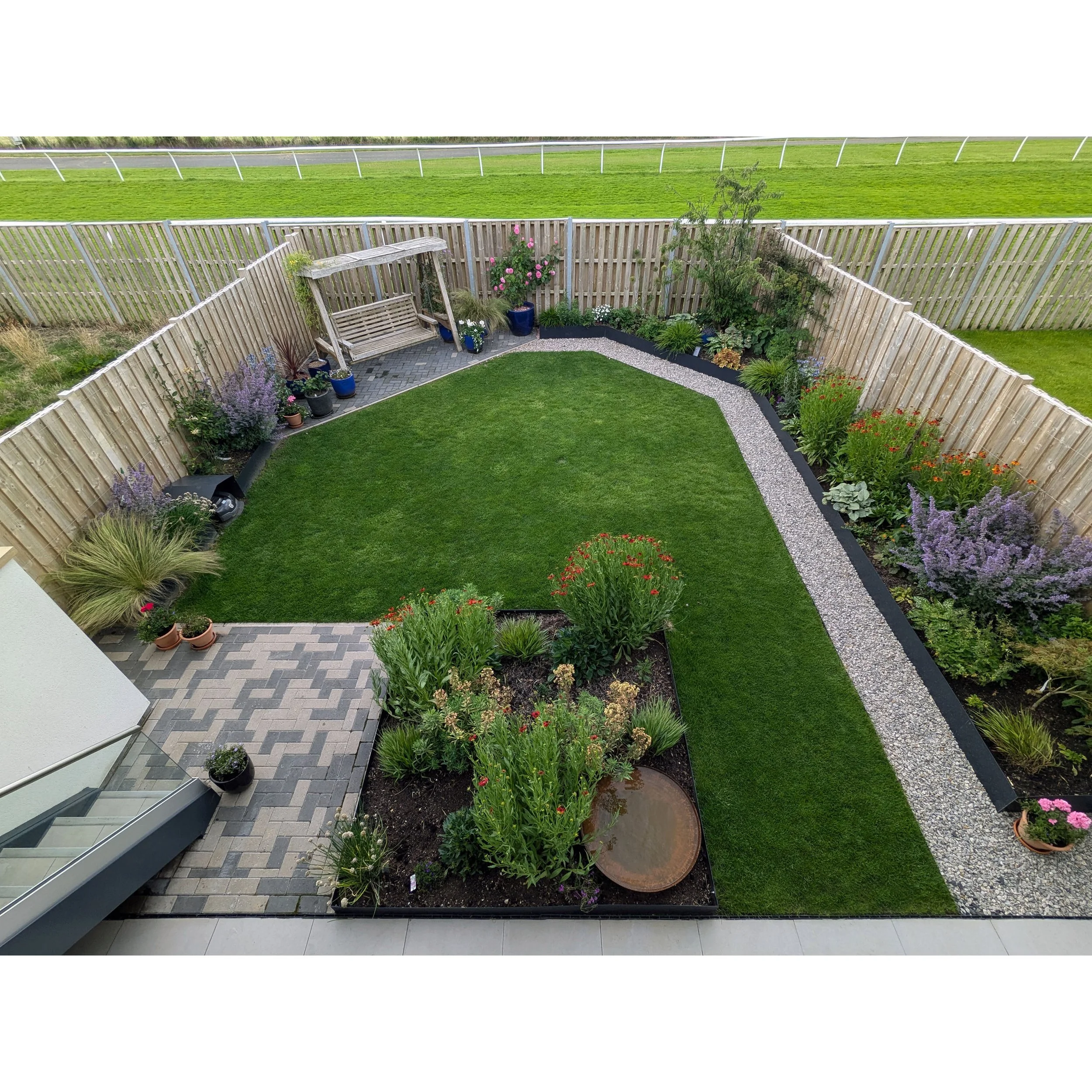 A contemporary garden with a lawn, raised metal flower beds, potted plants, a bench with a canopy, and a gravel pathway, fenced in and overlooking a racecourse.