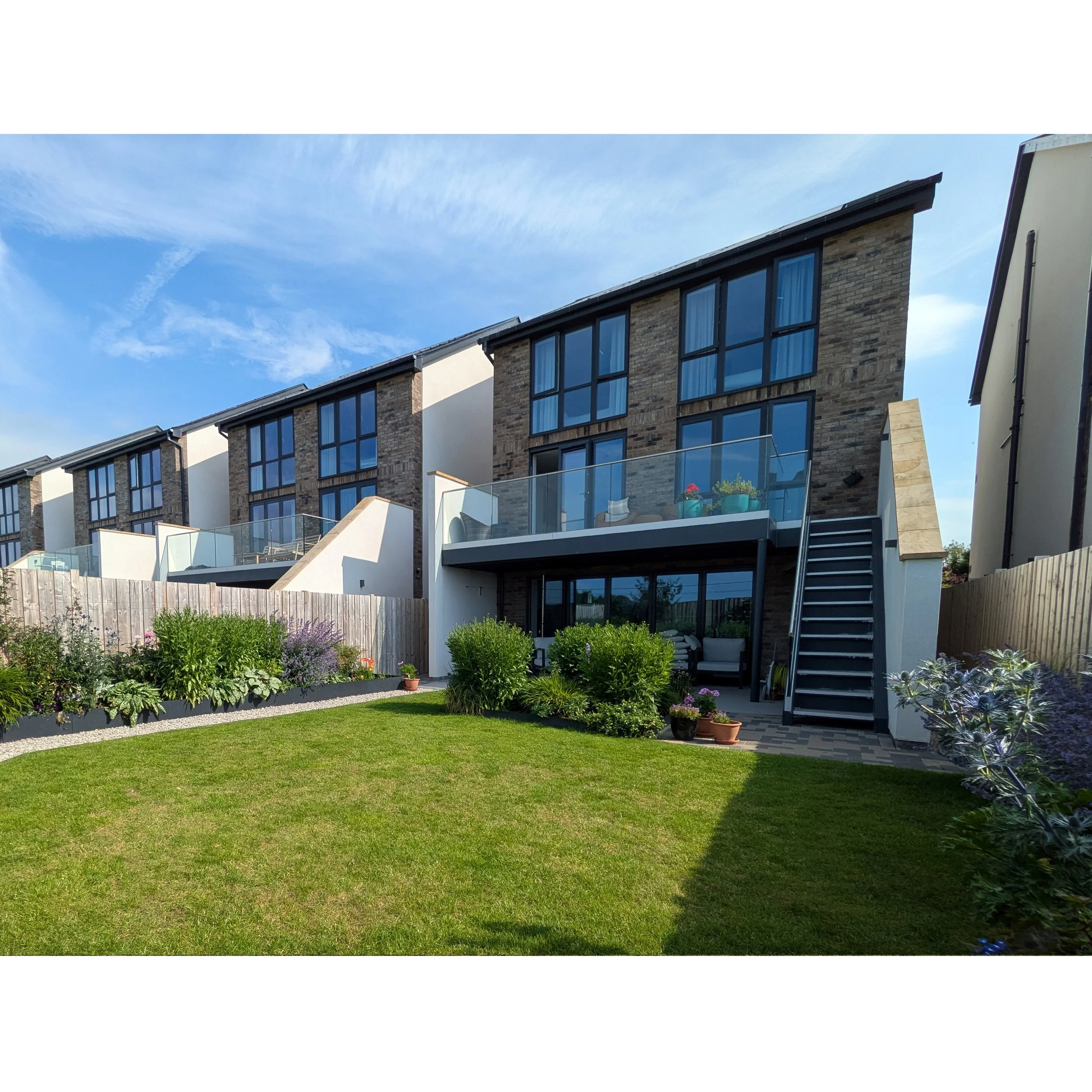 A contemporary townhouse with large windows and a cozy back garden with flowers, grass, and a small paved patio.