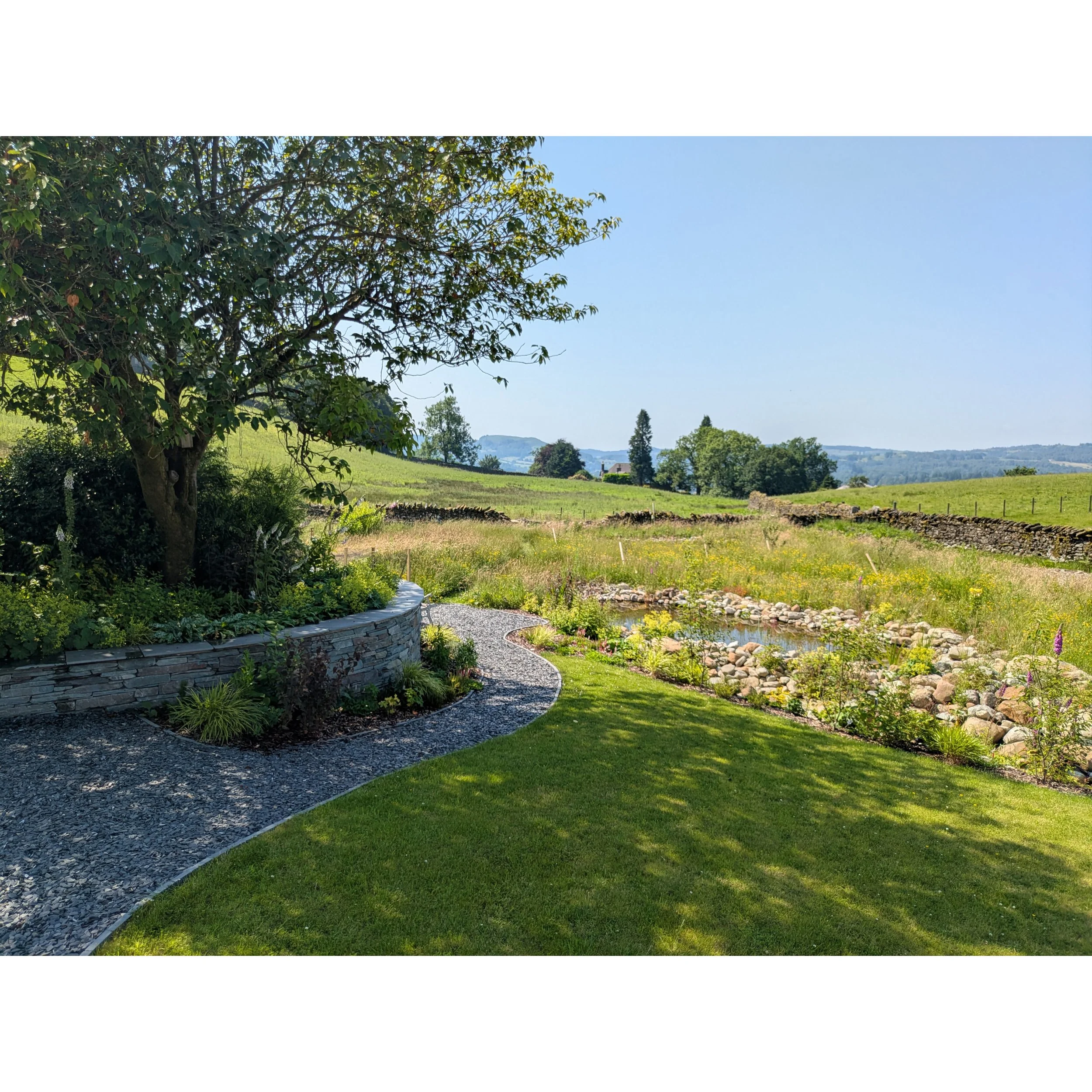 A scenic view of a garden with a wildlife pond in the foreground and rolling hills under a blue sky.