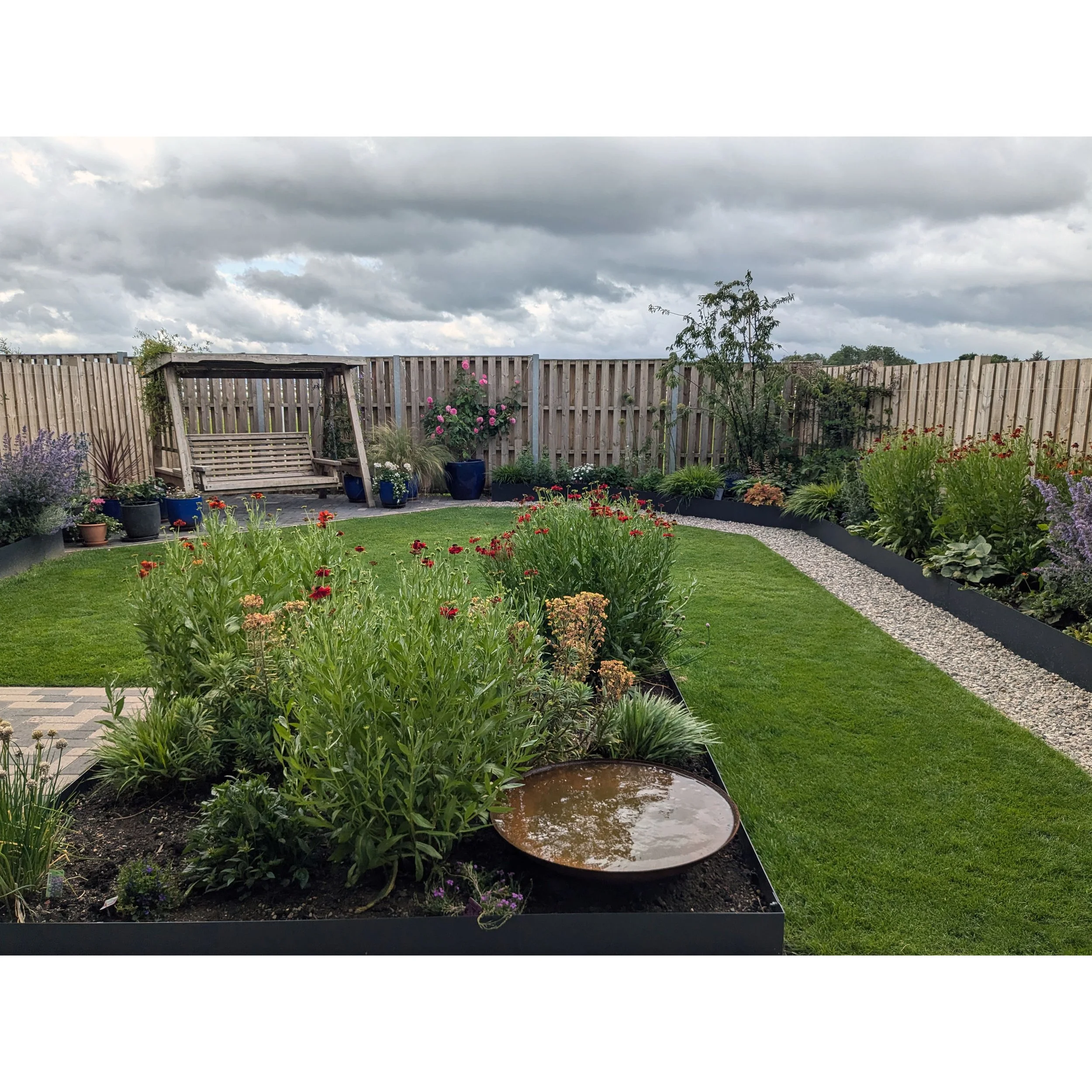 A contemporary back garden with a raised metal planters containing colourful flowers, a garden swing, potted plants, and a gravel pathway under a cloudy sky.