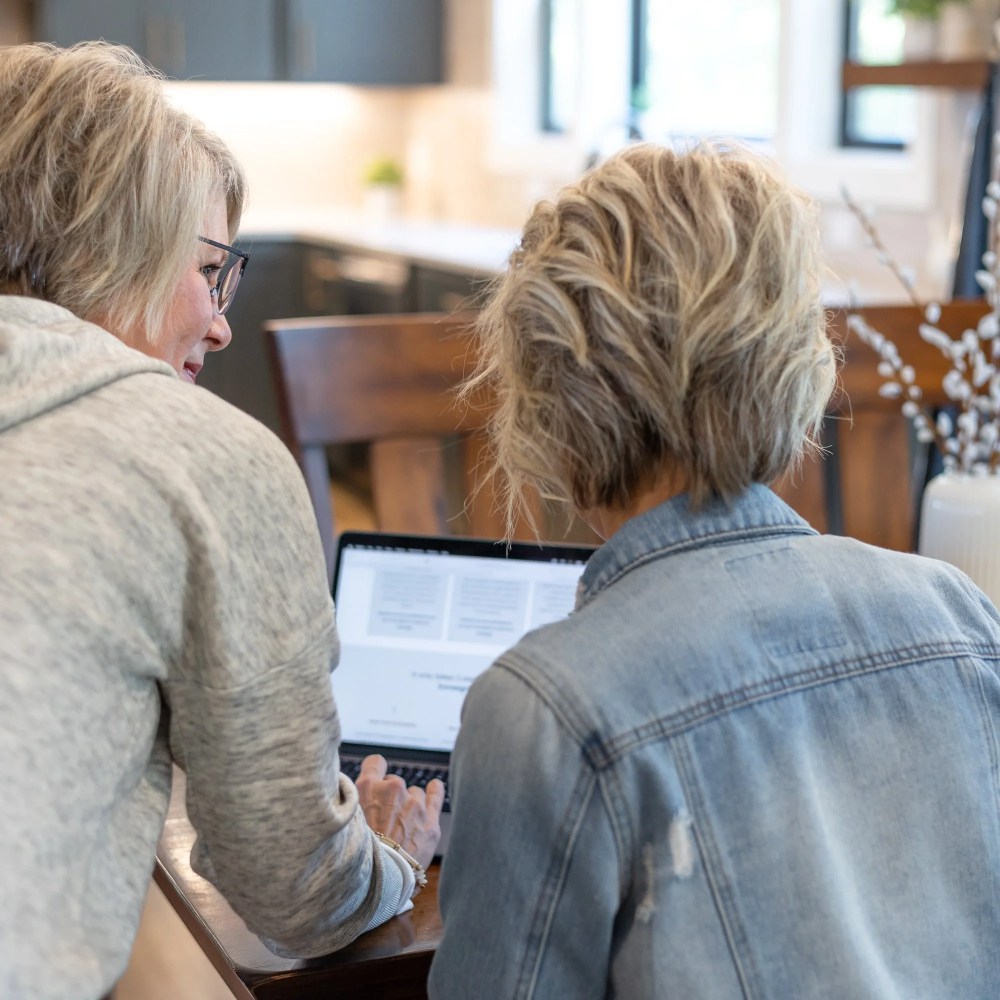 Two women with blond hair sitting at a wooden table, using a laptop in a bright room with large windows.