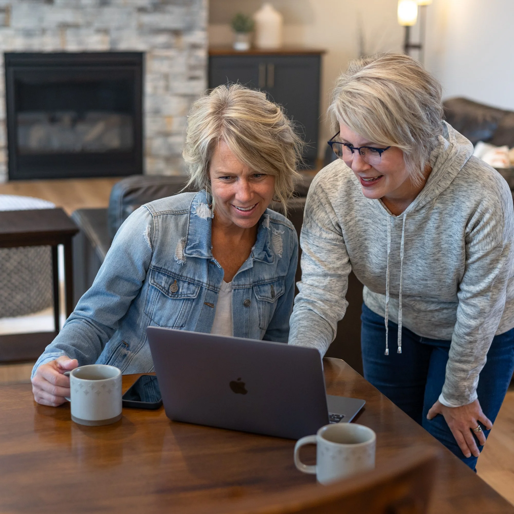 Heidi looking at a laptop computer working with a client and smiling in a cozy living room with a stone fireplace and a wooden table.