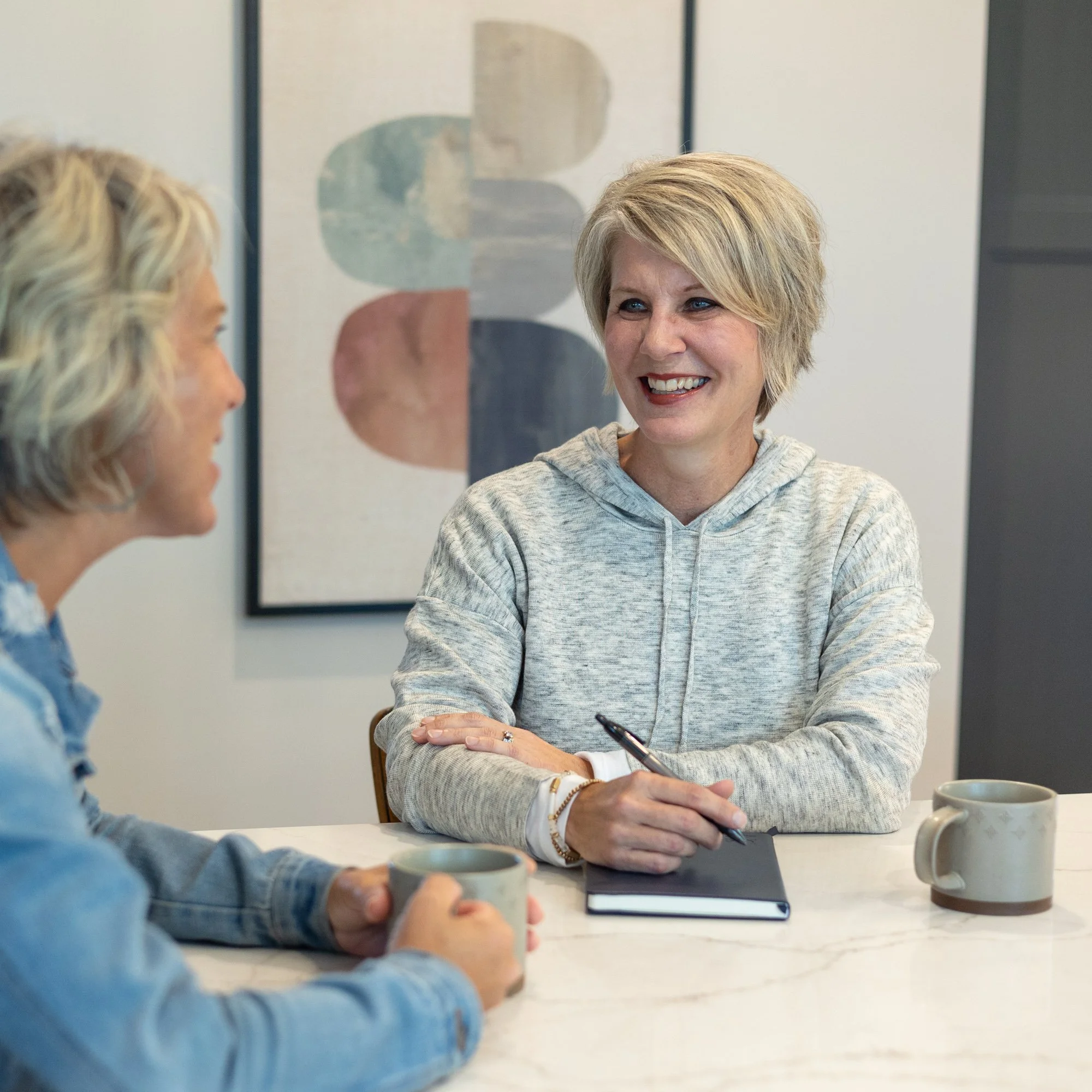 Two women sitting at a table in a casual setting, smiling and engaged in conversation. One woman has short blonde hair and the other has shoulder-length curly hair. They are holding mugs, and there are notebooks and a pen on the table.