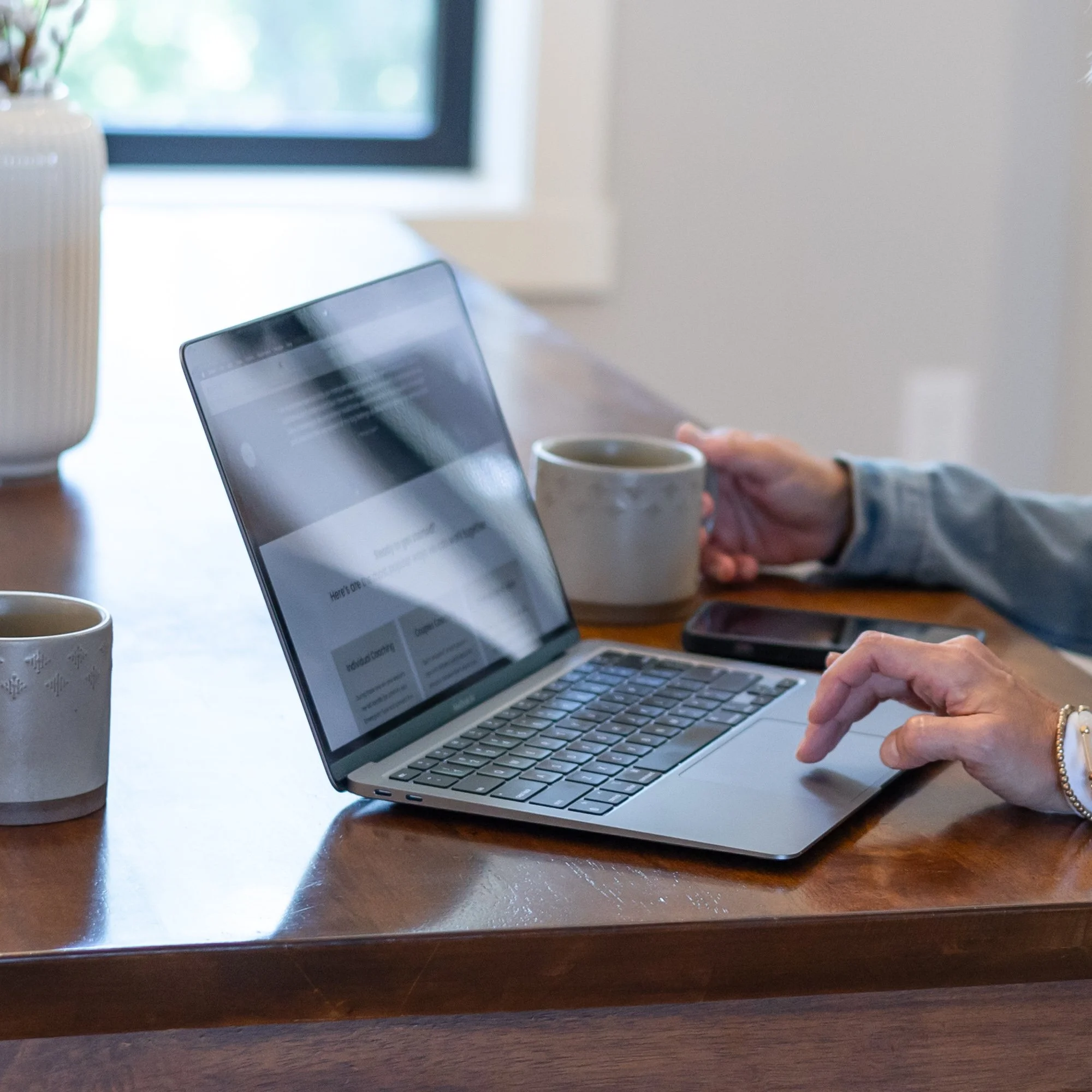 A person using a laptop computer at a wooden desk, with two cups of coffee and a ceramic vase with flowers nearby, against a background of a window.