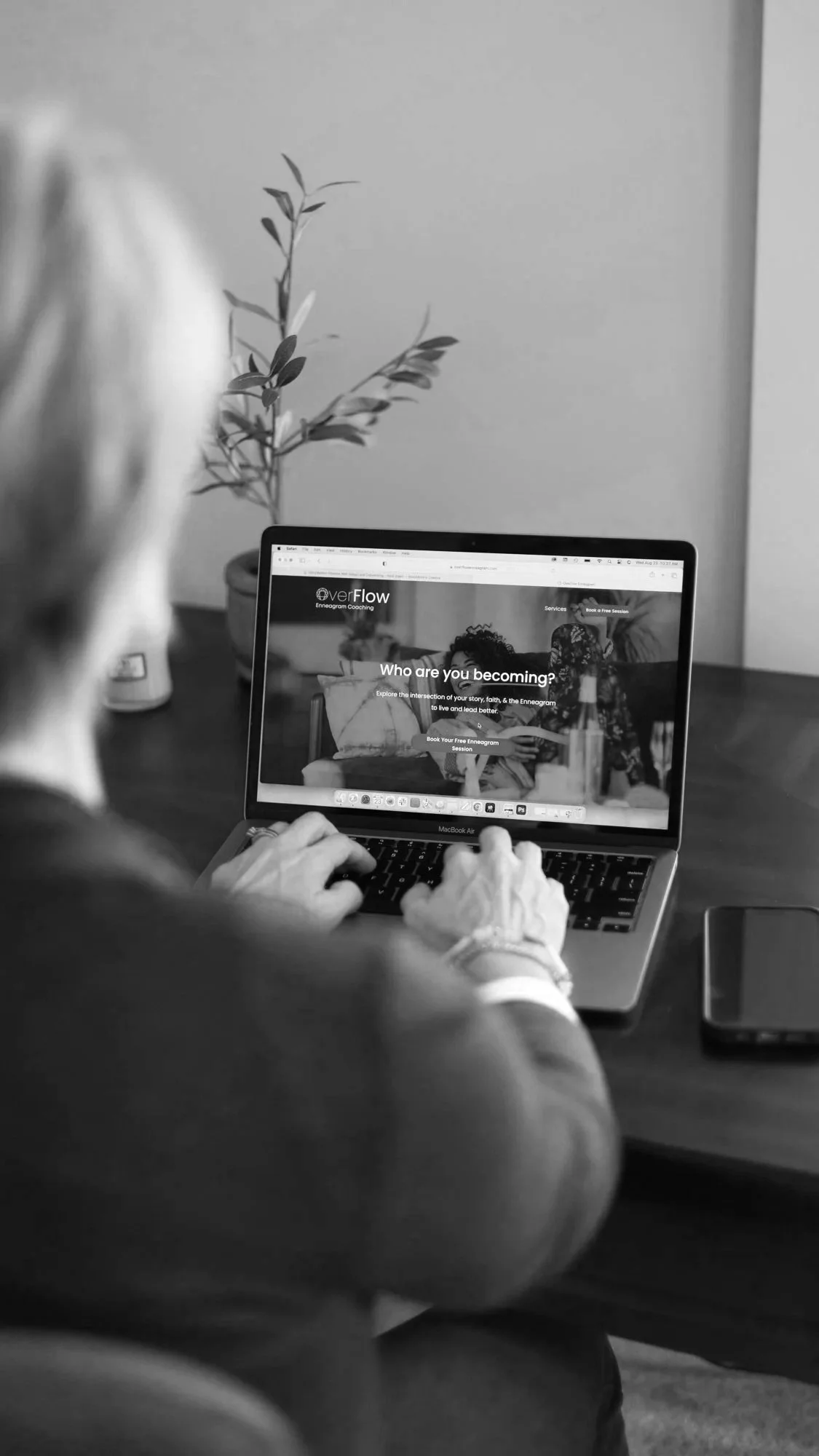 A person sitting at a desk using a MacBook Air laptop, viewing a webpage titled "Overflow Enneagram Coaching" with the question "Who are you becoming?" on the screen. There is a potted plant in the background, and a smartphone is placed on the desk.