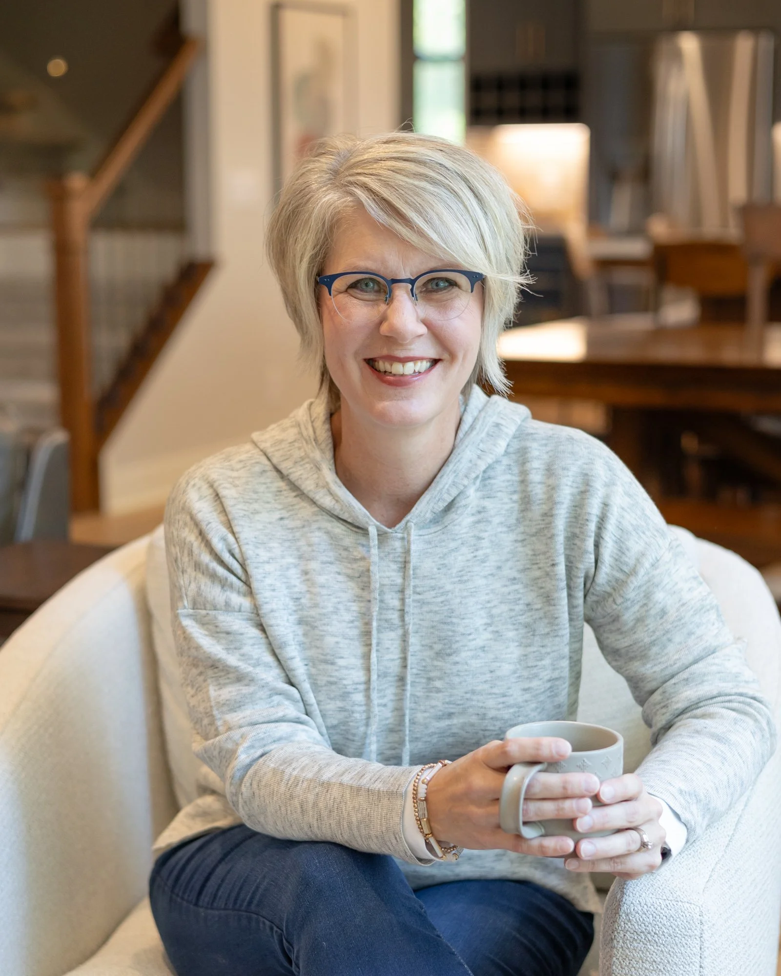 Heidi Zwart sitting on a beige chair holding a white ceramic mug, smiling at the camera in a cozy indoor setting.