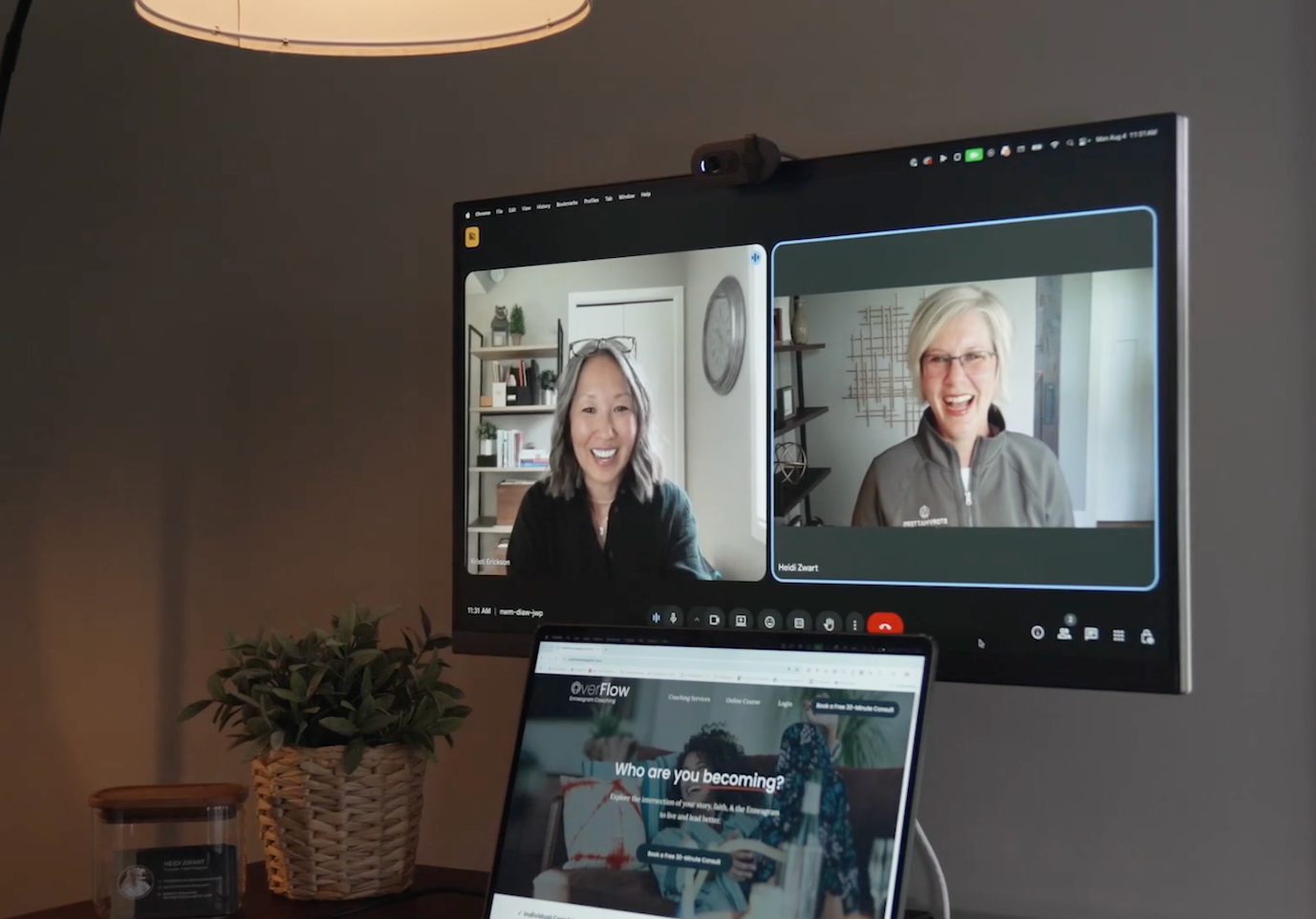 A video call on a computer screen shows two women talking, one with glasses and gray hair, the other with short blonde hair and glasses, both smiling. A laptop screen below displays a website called OverFlow with the question 'Who are you becoming?'. The desk has a potted plant in a woven basket and a candle.