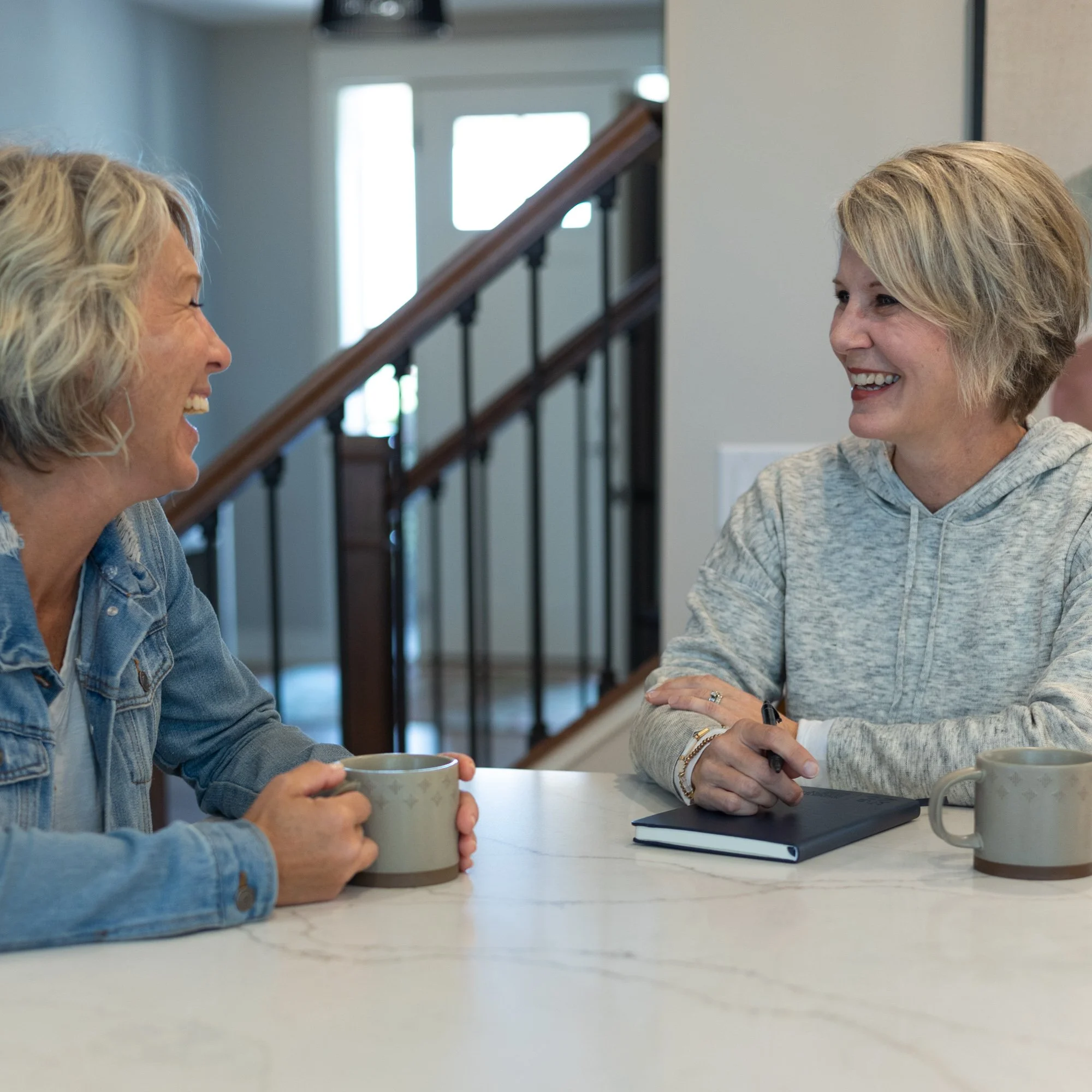 Heidi Zwart and a client having a conversation. One woman is holding a coffee mug, and the other has a notepad and pen. They are in a well-lit room with a staircase in the background.