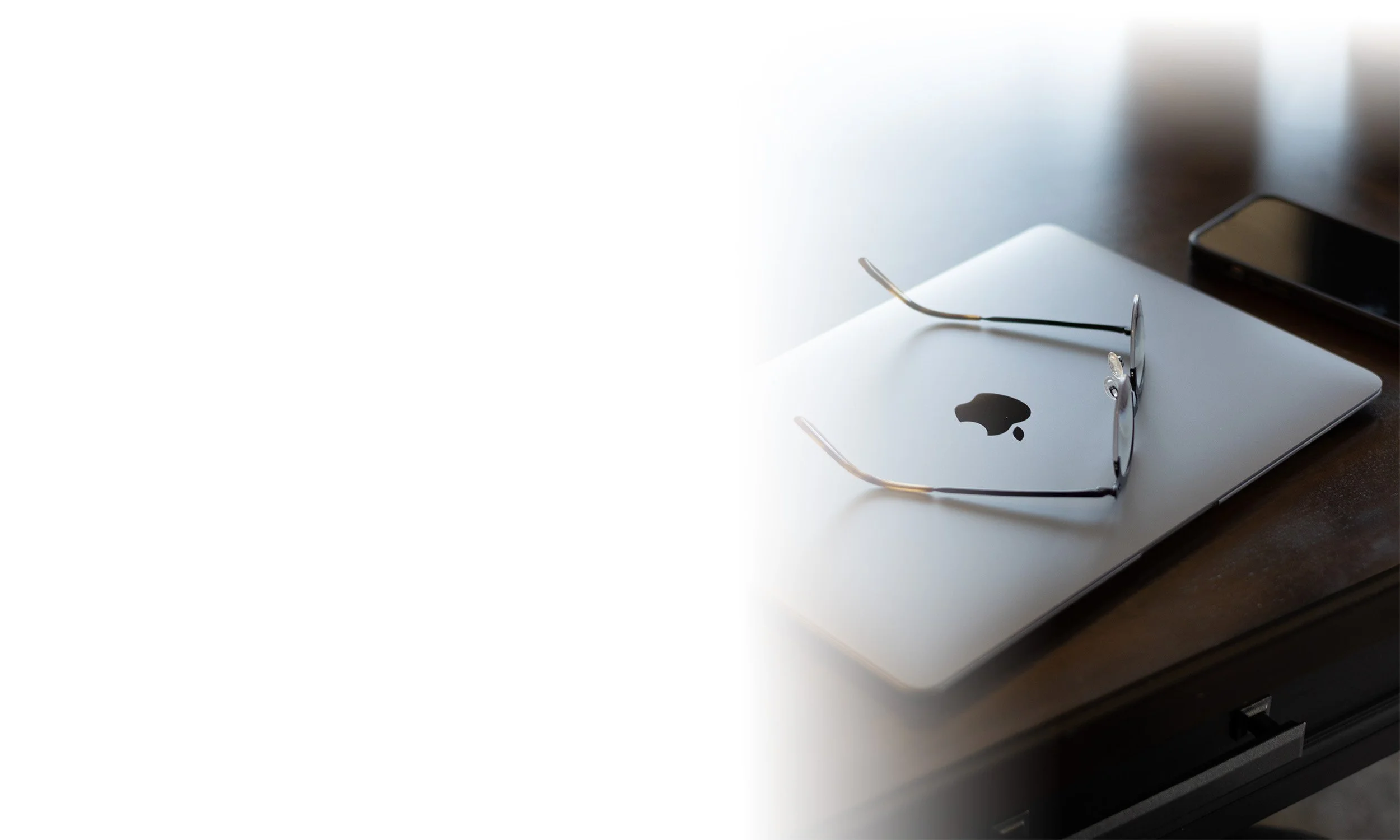 A closed Apple MacBook laptop with a pair of eyeglasses resting on top, and a smartphone placed nearby on a wooden surface.