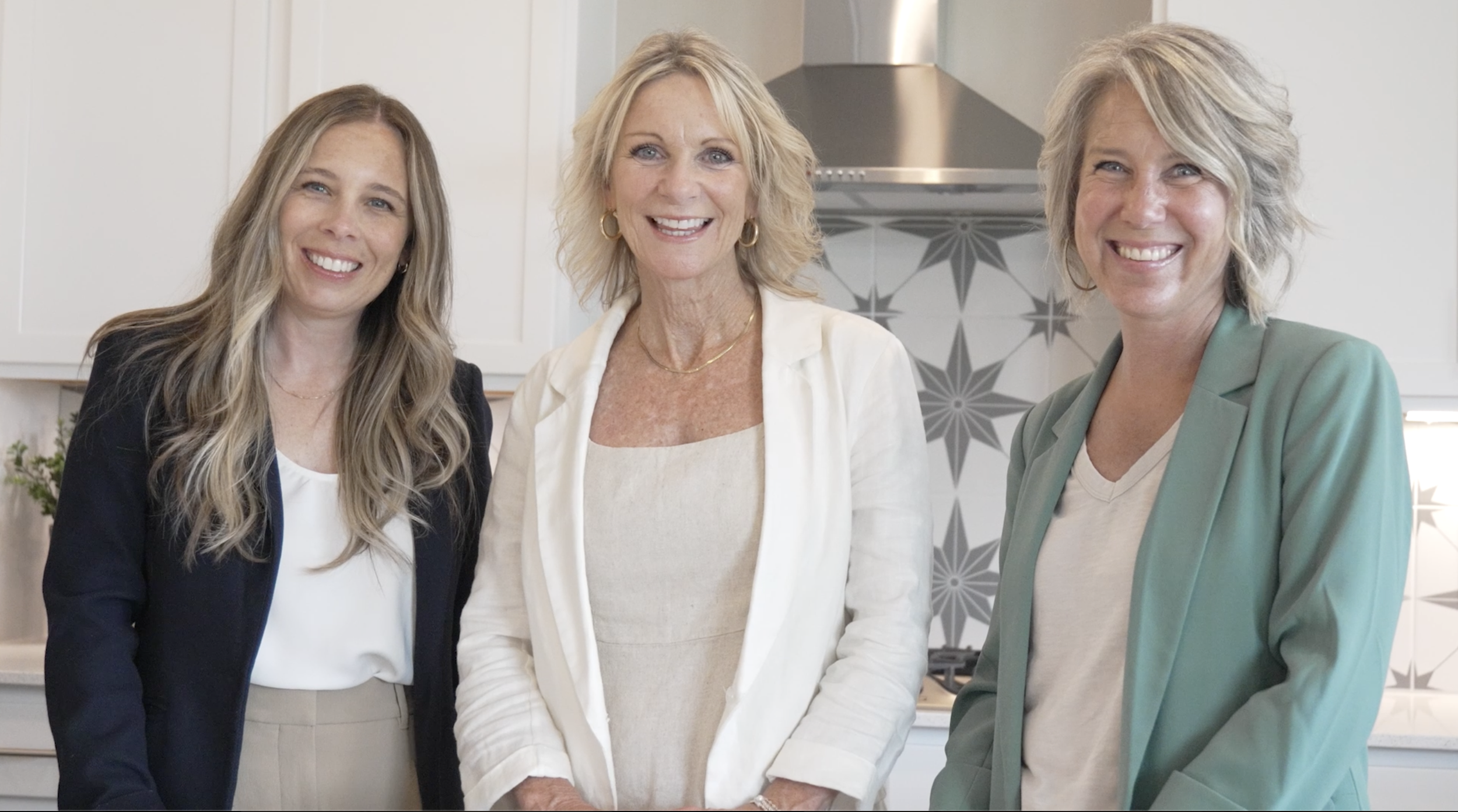 Three women smiling and standing close together in a kitchen, dressed in business casual attire.
