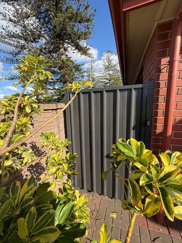 A gray Pedestrian gate attached to a brick house, surrounded by green plants and trees, with a bright blue sky in the background.