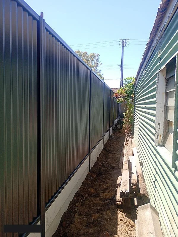 A narrow backyard space between a new black metal fence and an older green wooden house, with dirt on the ground and construction materials along the side.