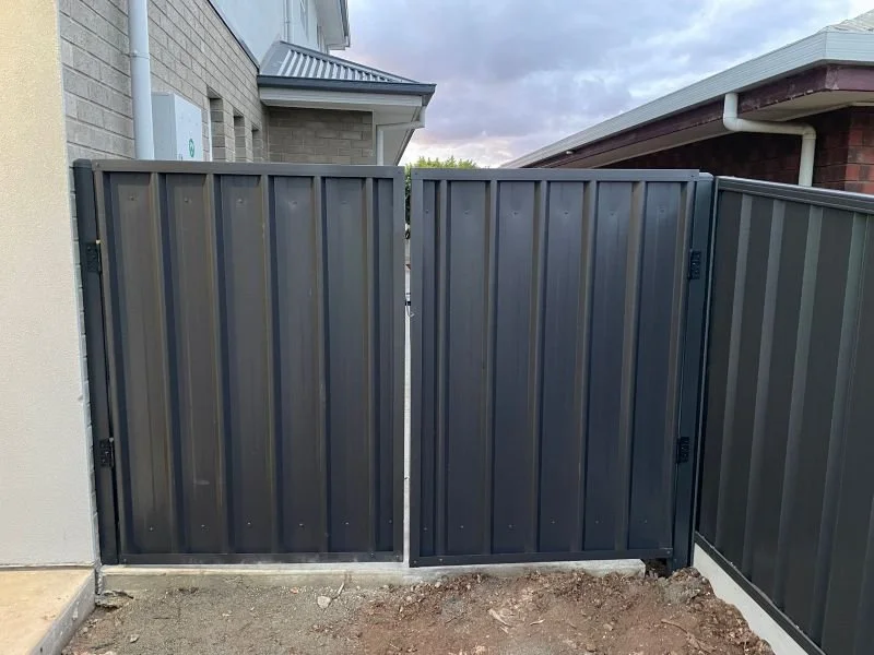 Black metal privacy gate with two sides, installed on a concrete base, next to a brick house and a neighboring house with a white roof.