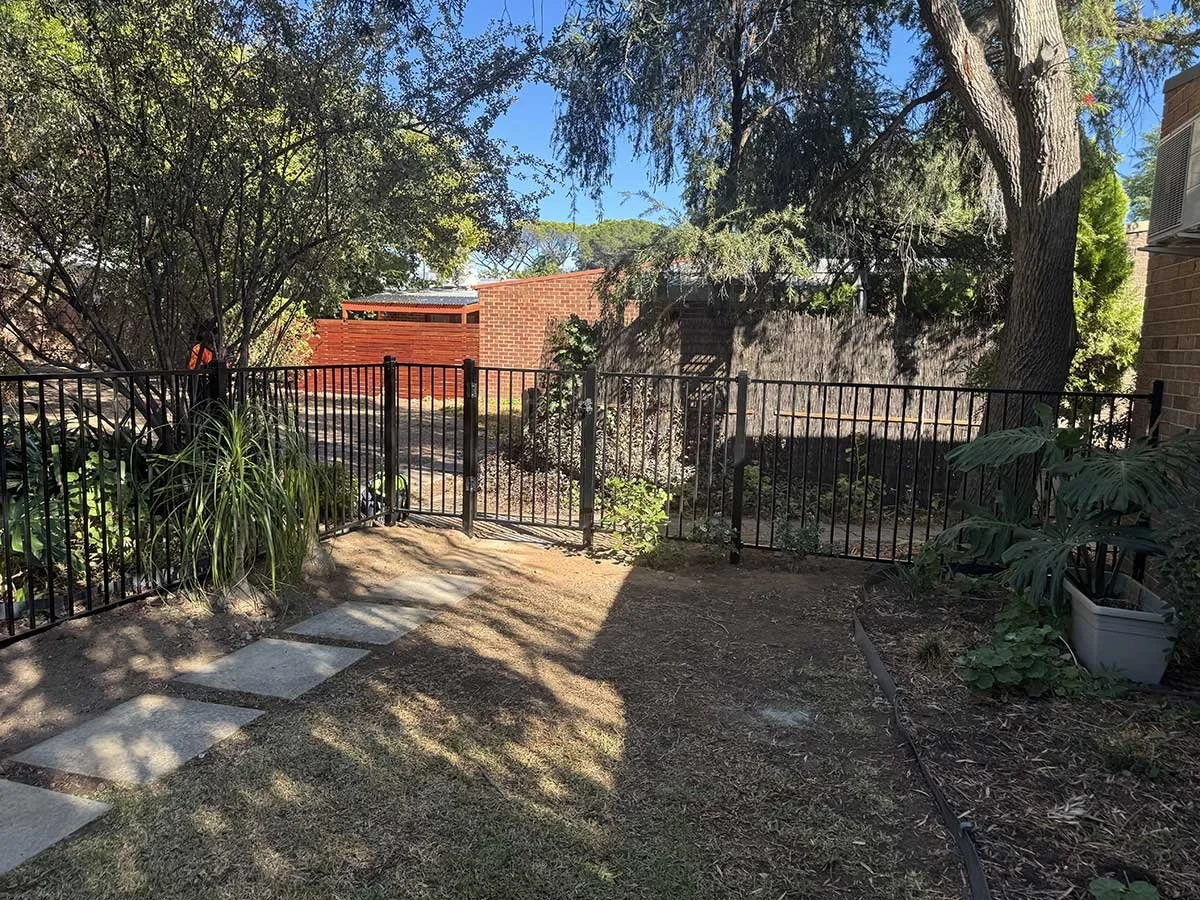 A backyard garden area with a black metal fence, stepping stones, plants, trees, and a brick building in the background, under a clear blue sky.