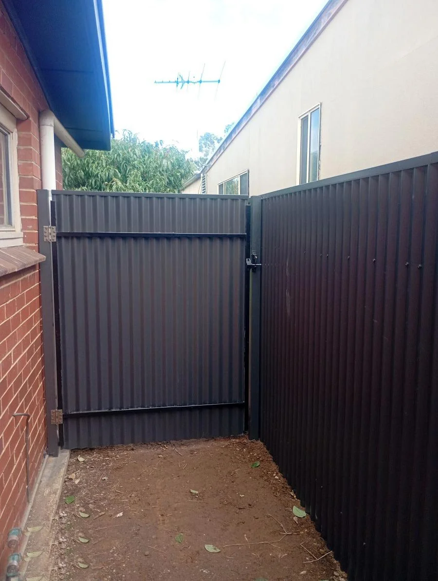View of a small enclosed backyard area with a dirt floor, surrounded by black metal fencing, and a black metal gate.