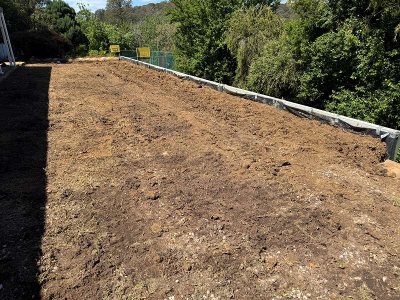 A cleared and leveled patch of dirt on a rooftop or elevated surface, enclosed by a guardrail, with trees and greenery in the background.
