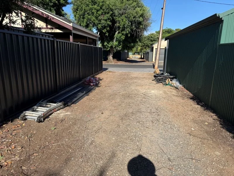 A dirt pathway between black metal fencing on the left and green fencing on the right. There are construction materials, including metal rods and black supplies, on both sides. In the background, there's a large tree and a narrow asphalt road leading