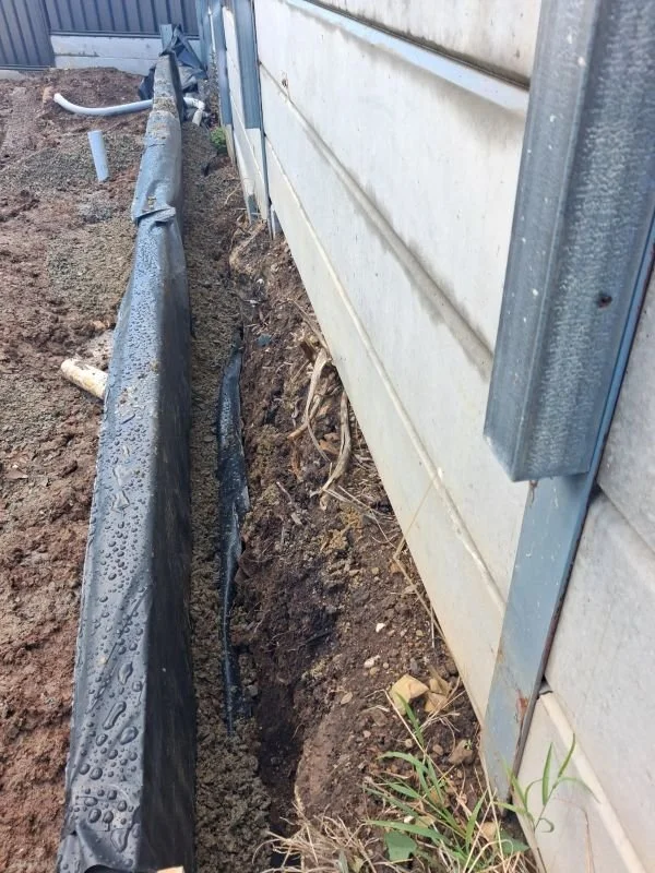 Excavation along the side of a house showing a trench with exposed pipes and a black water barrier or drain line, next to a house with vinyl siding.