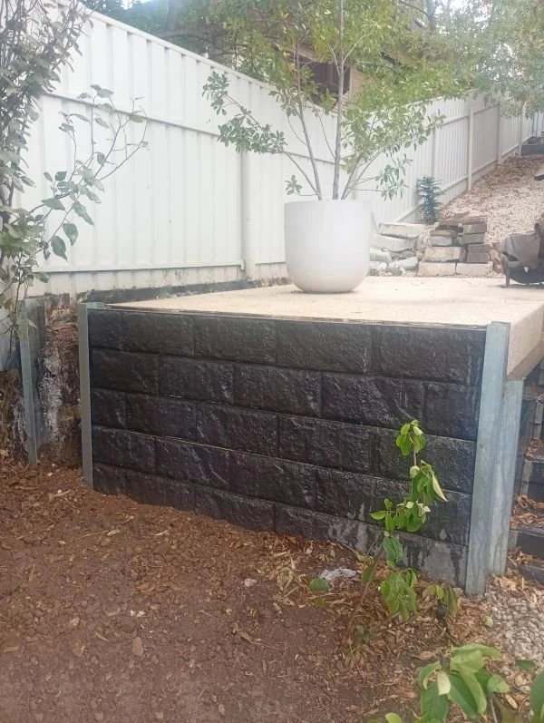 Part of a backyard with a black painted brick wall, a small plant in front, and a large white pot with a tree on a concrete platform. A white fence and some patio furniture are in the background.