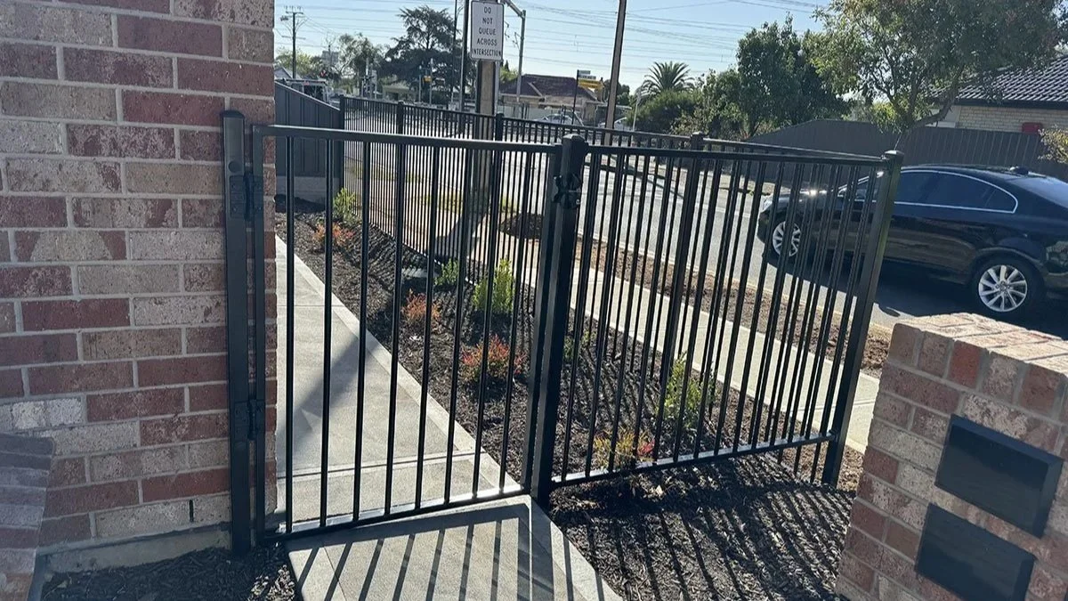 A black metal gate and fence enclosing a sidewalk with small plants, with a brick wall on either side, in front of a street with cars and trees under a clear sky.