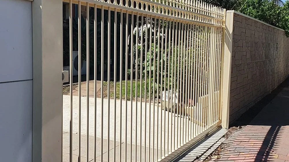 A cream-colored metal gate with vertical bars and decorative top spikes, leading to a driveway and garden area, with a stone wall on the right side and a sidewalk on the left.