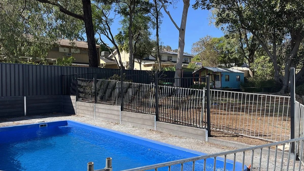 A small in-ground pool enclosed by a tubular fence, with trees and a house in the background under a clear blue sky.