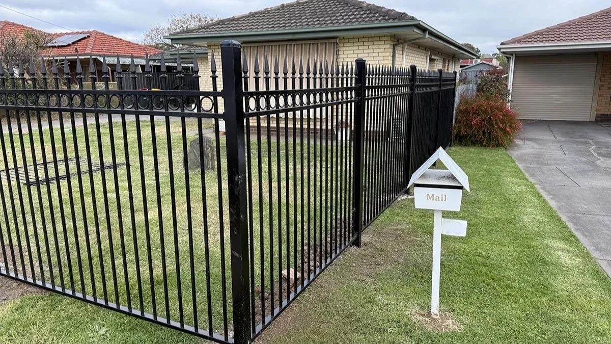 A black metal fence surrounds a front yard with a small gravestone visible inside. A white mailbox labeled 'Mail' is mounted on a pole near the sidewalk. Residential houses with brick and tiled roofs are in the background under a cloudy sky.