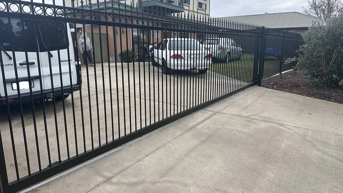 A black metal tubular fence with pointed tips, enclosing a driveway with parked cars in front of a two-story house, with a balcony and a lawn on the side.