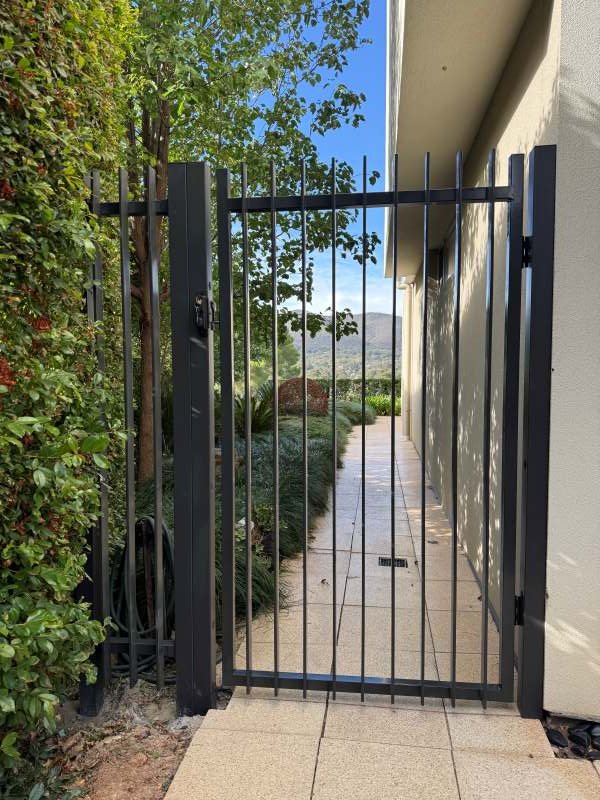 Black metal gate with vertical bars at the entrance of a pathway, with greenery on the left and a house wall on the right, leading to a scenic view of trees and hills in the distance under a blue sky.
