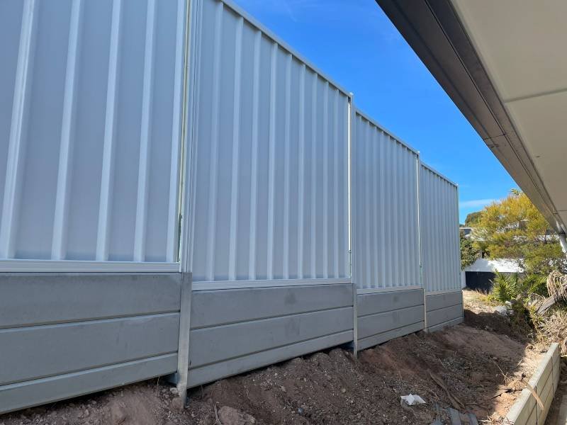 A close-up view of a tall, white metal building wall with vertical panels, part of a construction site with dirt and a few small plants nearby, under a clear blue sky.