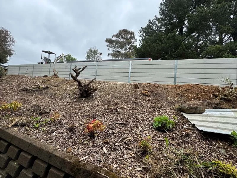 A backyard scene with a sloped soil area, a few small plants, and a large metal sheet partially buried. A gray fence runs along the top of the slope, with trees and an overcast sky in the background. There is a construction worker and some equipment 