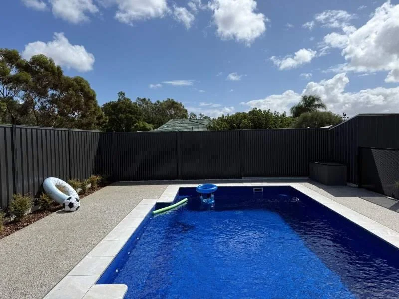 Backyard with a swimming pool, pool float, and inflatable soccer ball, surrounded by a black fence under a bright blue sky with scattered clouds.