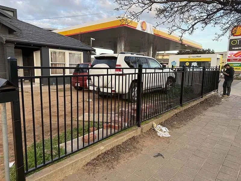 A Shell gas station with vehicles parked in front, surrounded by a black metal fence, with a person standing on the sidewalk using their phone.
