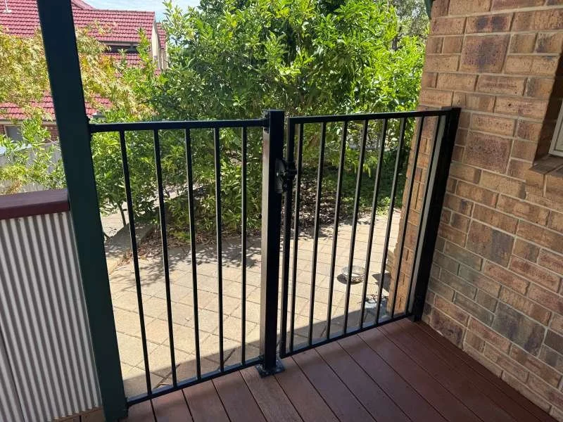 Black metal gate on a patio with brick wall on the right and green trees in the background.