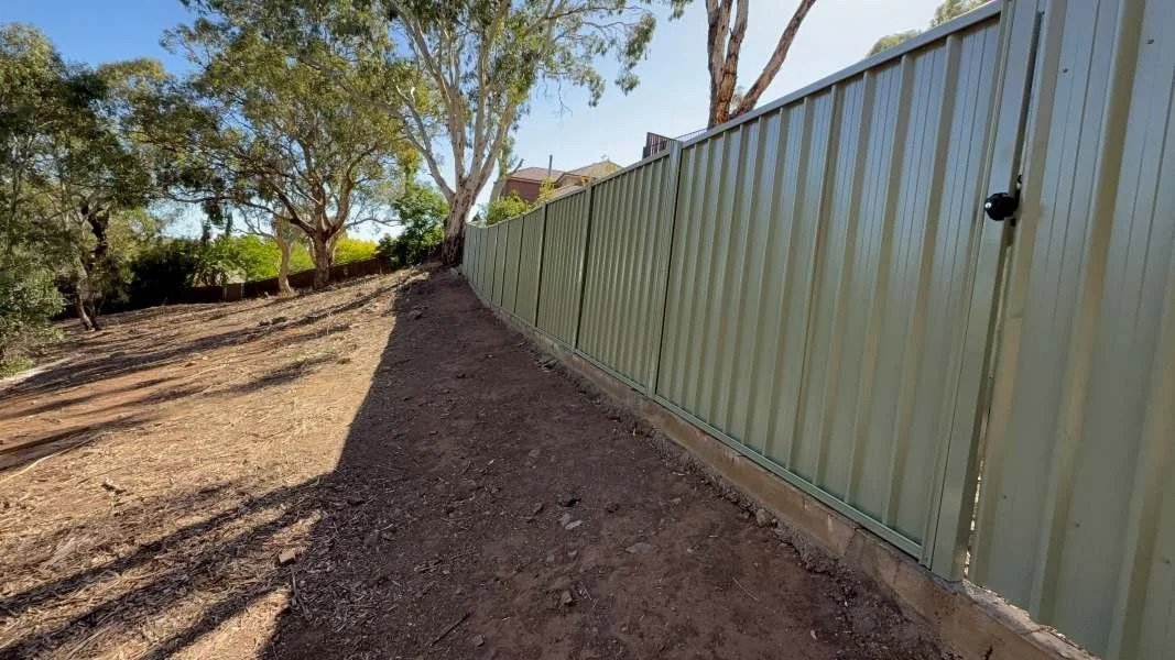 A landscaped yard with trees and a green metal fence along a sloped dirt terrain on a sunny day.