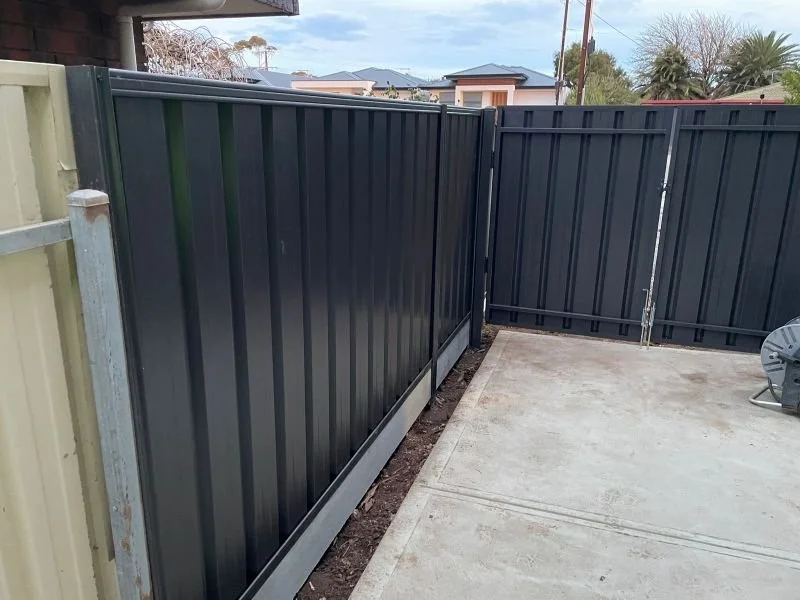 A newly installed dark gray metal fence surrounds a small outdoor area with concrete flooring, adjacent to a yellow and green fence. The sky is partly cloudy and neighboring houses with trees are visible in the background.