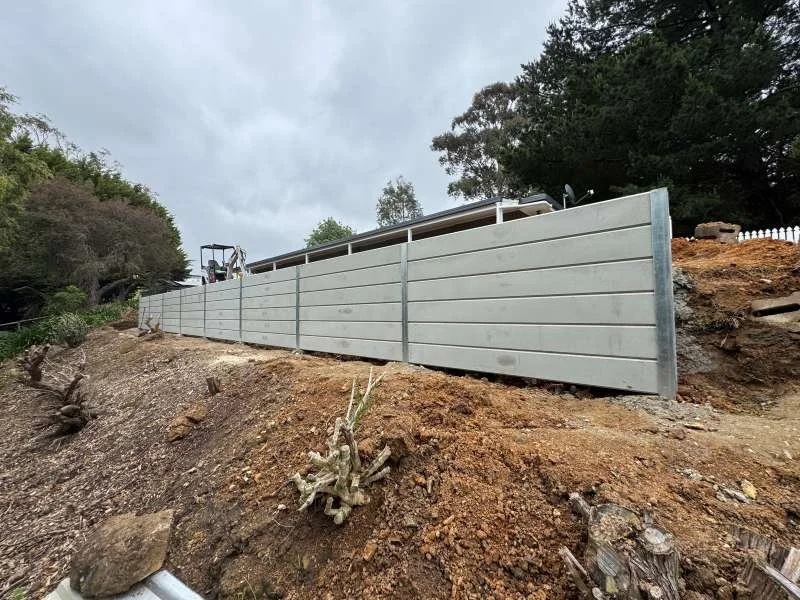A long metal retaining wall on a hillside, with trees and cloudy sky in the background.