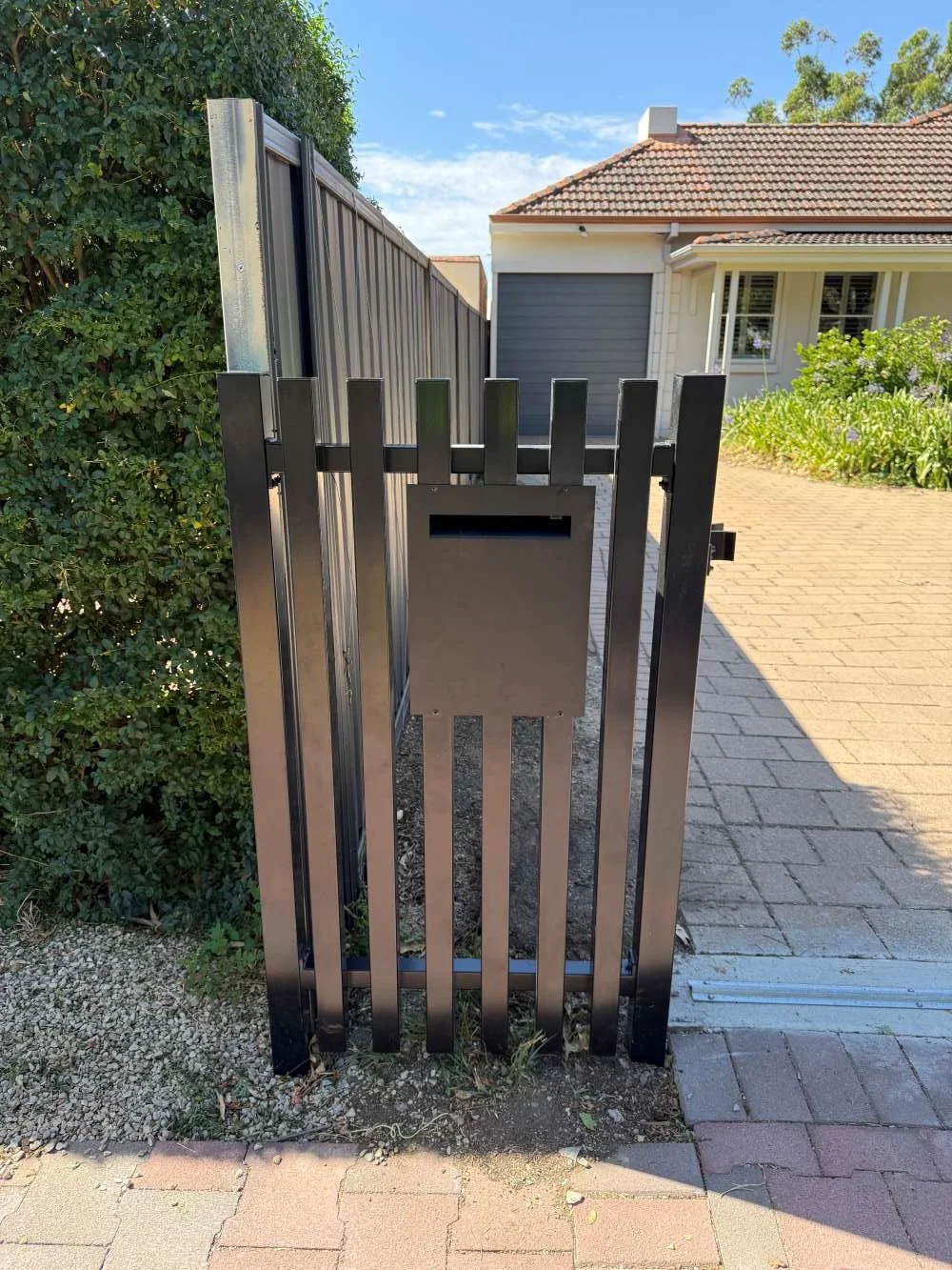 Black metal gate with vertical slats, attached to a hedge on the left and a house with a garage and window in the background.