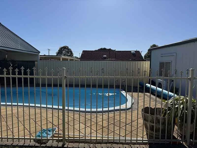 In-ground backyard swimming pool enclosed by a white metal fence, with a tiled deck area, a shed, and a large blue pool cover rolled up on the side. The sky is clear and blue.