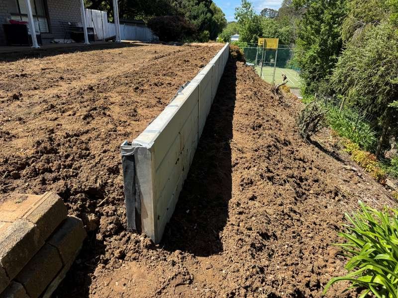 A construction site with a retaining wall being built along a sloped hillside, surrounded by dirt and greenery, with a tennis court visible in the background.