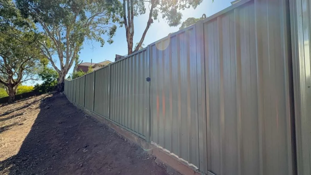 Metal fence running along a dirt slope with trees in the background under a blue sky.
