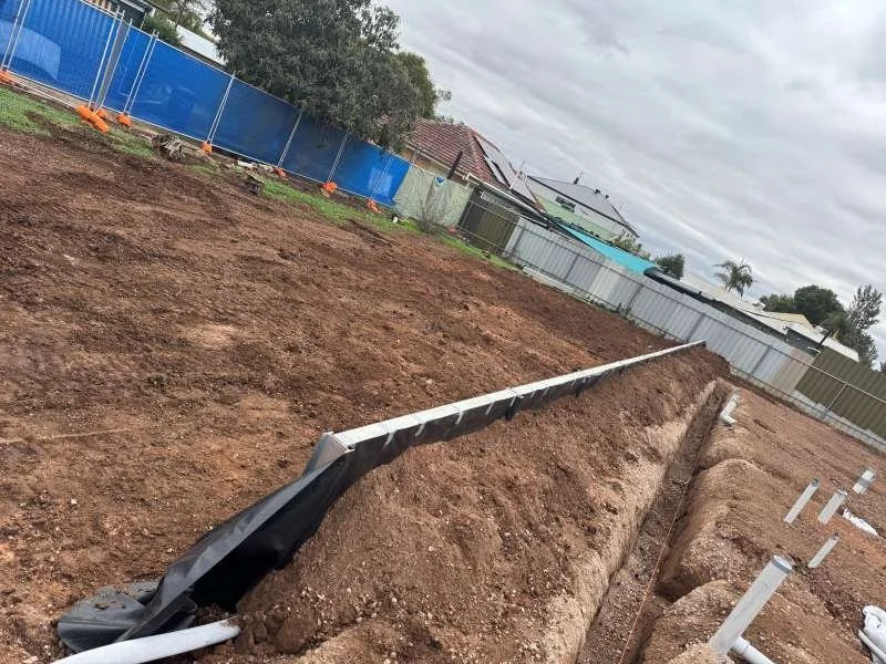 A construction site with a trench and a long black pipe lying on the dirt, surrounded by fences and residential houses in the background under a cloudy sky.