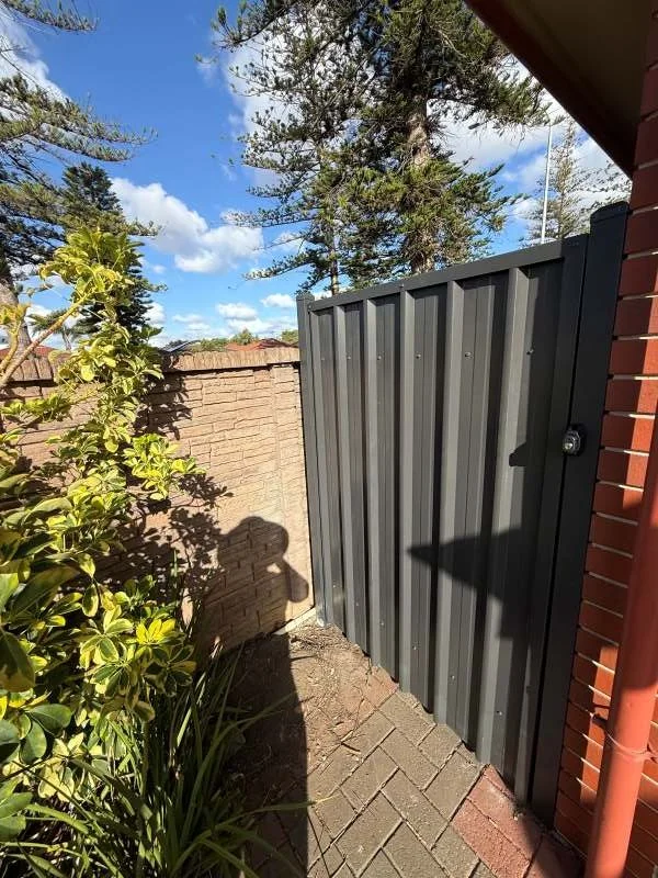 Small backyard corner with a dark gray metal gate, a brick wall, green plants, and tall trees in the background under a blue sky with clouds.