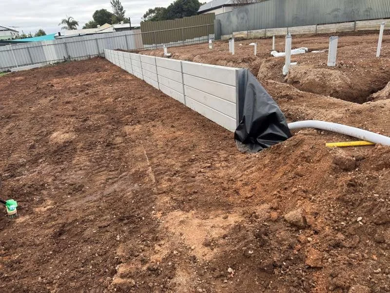 Construction site with a newly built white retaining wall on a patch of excavated dirt, piping and fencing in the background.